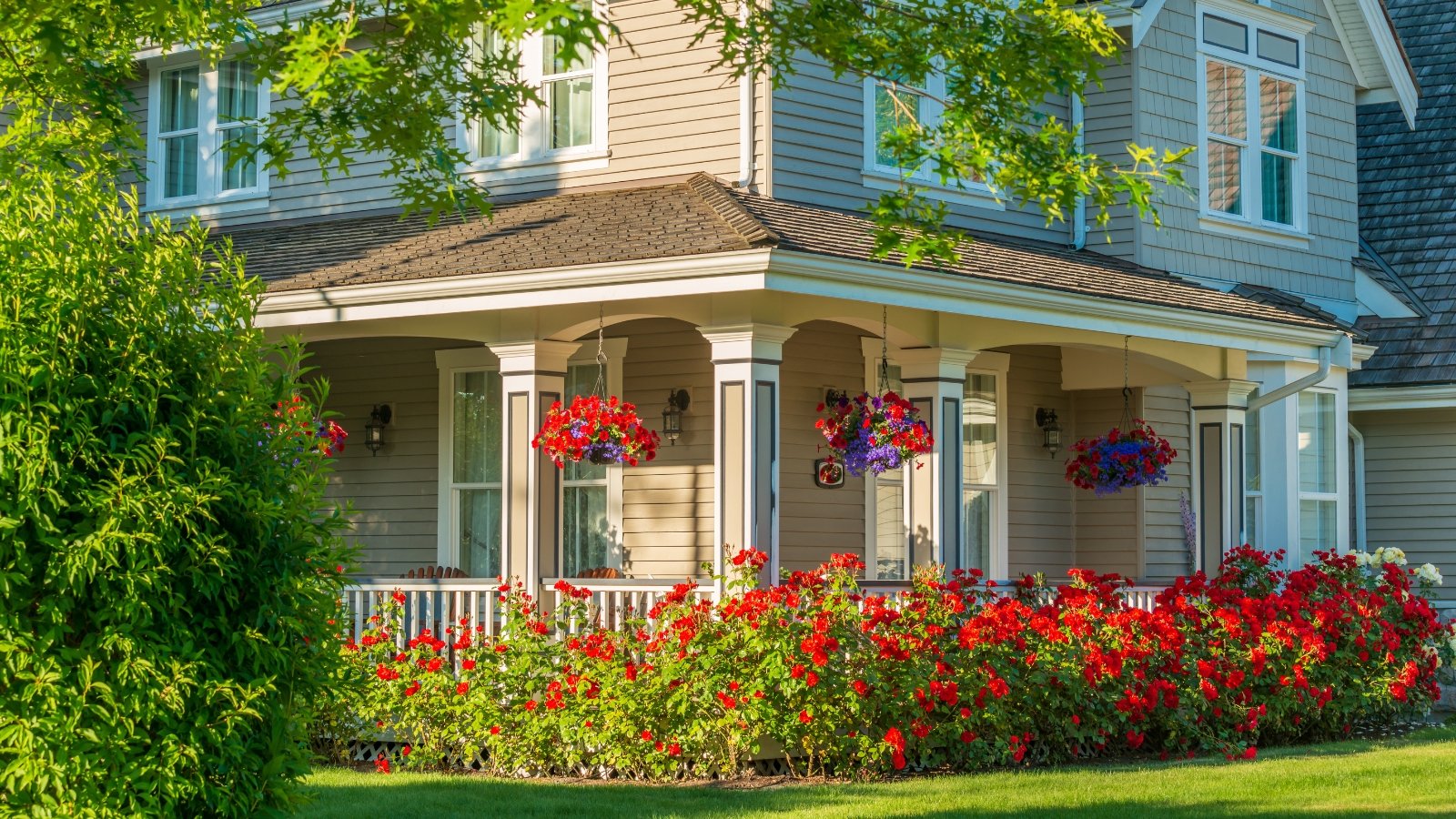 A gorgeous large soft blue house is surrounded by a green lawn, brightly blooming flower beds, tall trees, and bushes, with its porch decorated by hanging baskets of red and purple flowers, showcasing easy landscaping tweaks that enhance curb appeal.