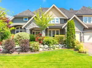 A large classic house with triangular roofs and wide white windows is complemented by a front yard featuring stone-edged flower beds filled with evergreen and flowering shrubs, tall and low trees, and a freshly mown green lawn, offering inspiration on how to avoid the most common landscaping mistakes.