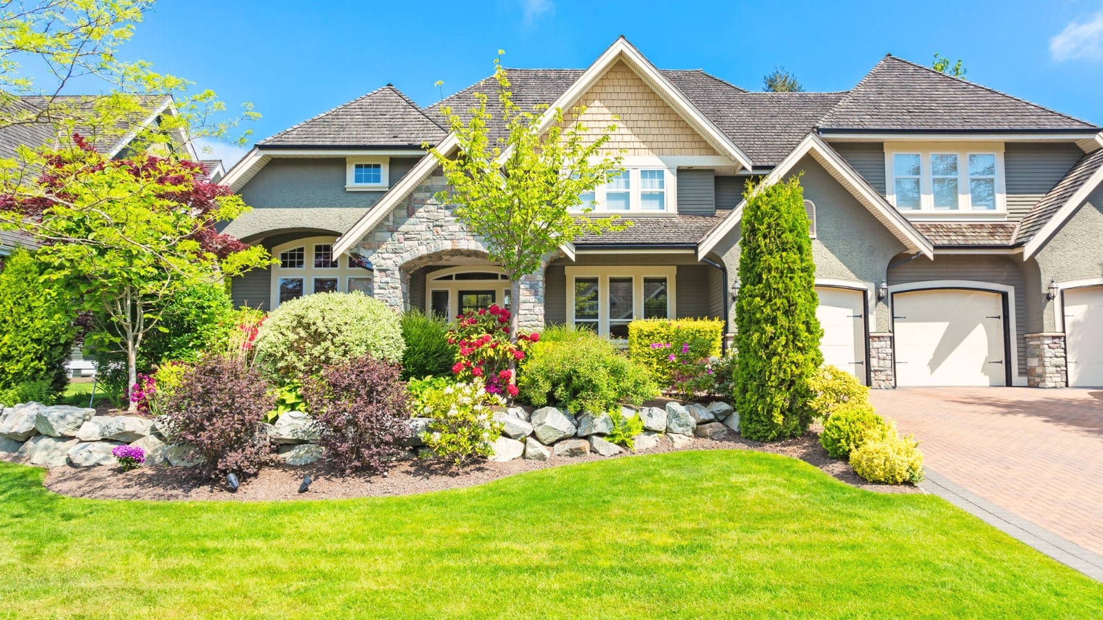 A large classic house with triangular roofs and wide white windows is complemented by a front yard featuring stone-edged flower beds filled with evergreen and flowering shrubs, tall and low trees, and a freshly mown green lawn, offering inspiration on how to avoid the most common landscaping mistakes.