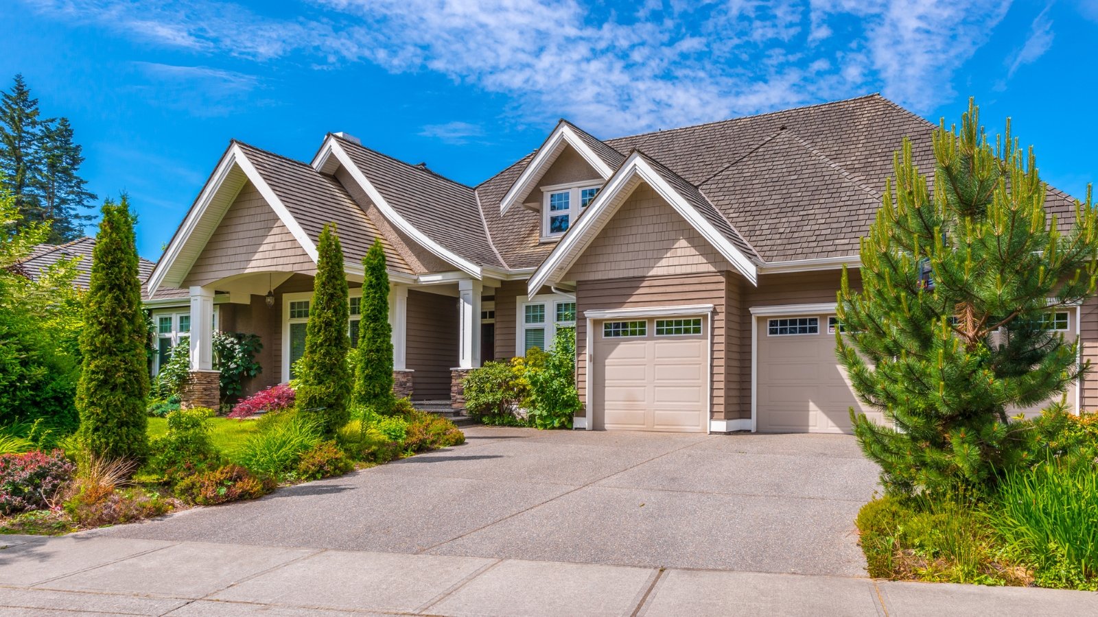 A luxurious large house with triangular roofs and white window frames is surrounded by a neat, well-landscaped front yard with evergreen trees, perennial shrubs, groundcover plants and ornamental grasses.