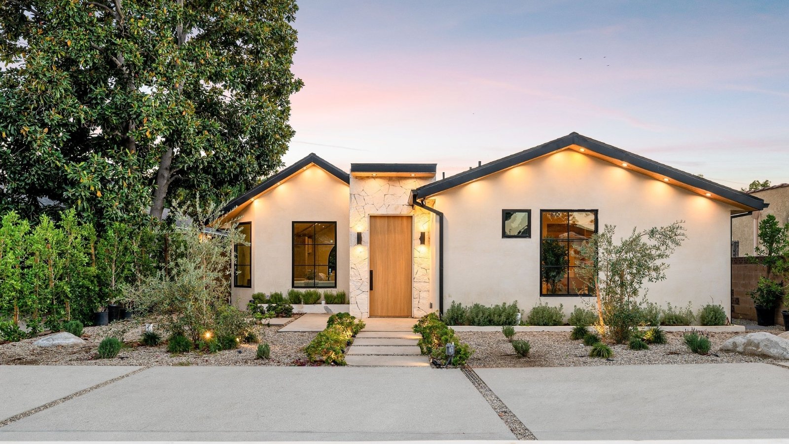 A modern, stylish home illuminated by lanterns with a minimalist front garden featuring xeric plants and trees set in mulched pebble soil.