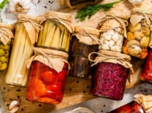 Glass jars filled with corn, peppers, beets, garlic, olives, and mushrooms, a way to preserve garden harvest for later use.