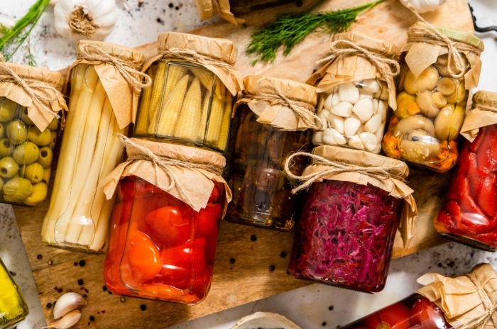 Glass jars filled with corn, peppers, beets, garlic, olives, and mushrooms, a way to preserve garden harvest for later use.