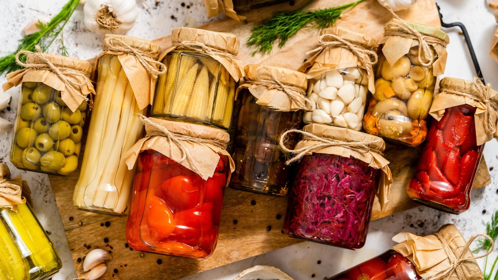 Glass jars filled with corn, peppers, beets, garlic, olives, and mushrooms, a way to preserve garden harvest for later use.