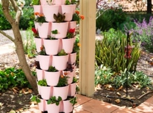 Various types of greenery and vibrant flowering plants flourish in a soft pink GreenStalk Planter, showcasing the beauty of vertical gardening systems in a sunny, blooming garden.