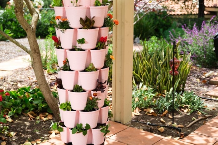 Various types of greenery and vibrant flowering plants flourish in a soft pink GreenStalk Planter, showcasing the beauty of vertical gardening systems in a sunny, blooming garden.