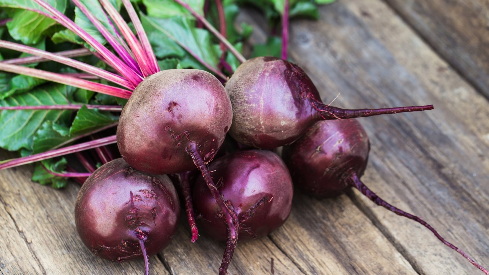 Round, deep red beet roots rest on a wooden surface, with upright, sturdy green stems and broad, slightly crinkled leaves in shades of green and reddish veins. 