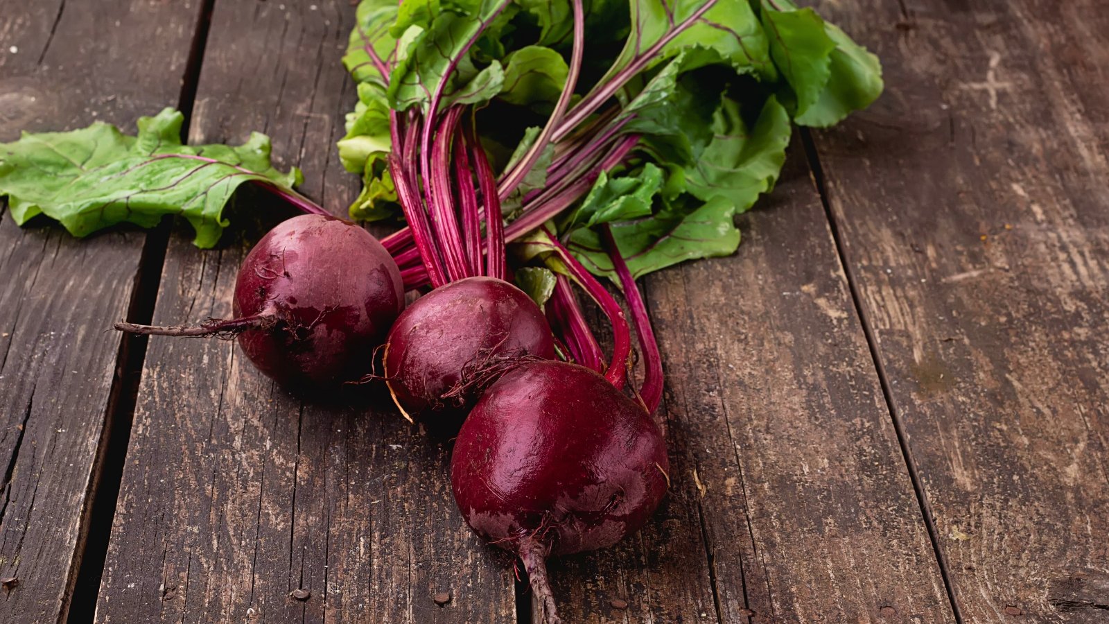 A bunch of freshly harvested beets with deep red, round roots and vibrant green leaves with reddish veins rests on a wooden surface.