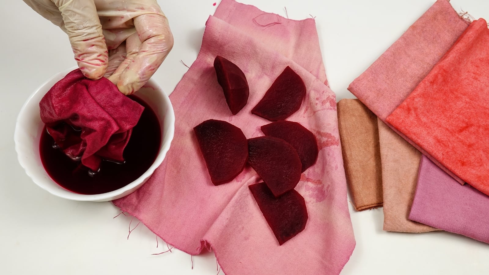 A cotton fabric sample is being dyed in beet broth with sliced beets placed on the red-toned cloth beside a bowl of the deep-colored liquid.