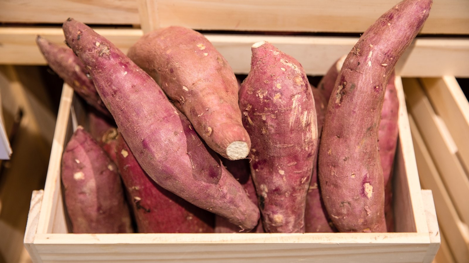 Cured sweet potato tubers with oval to slightly oblong shapes and pinkish skin fill a wooden box, their surfaces smooth and clean.