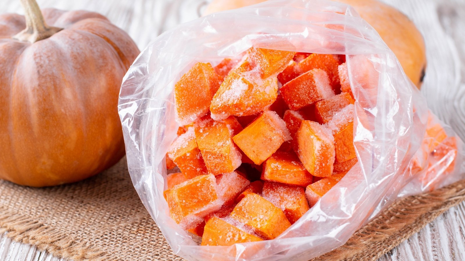 Pieces of frozen bright orange pumpkin are packed in a clear plastic bag on a table beside ripe, fresh whole pumpkins.