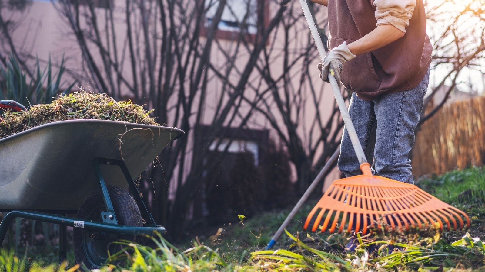 Gardener in gloves and a hoodie raking garden debris next to a cart filled with branches and leaves.