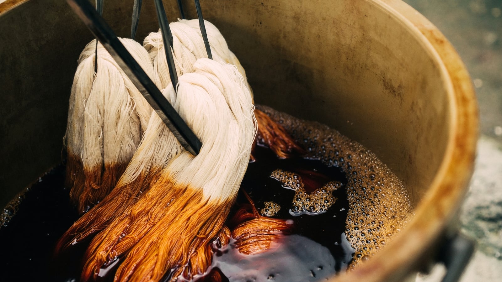 Cotton fabric tied to sticks hangs over a large pot of brown-orange dye water during a natural dyeing process.