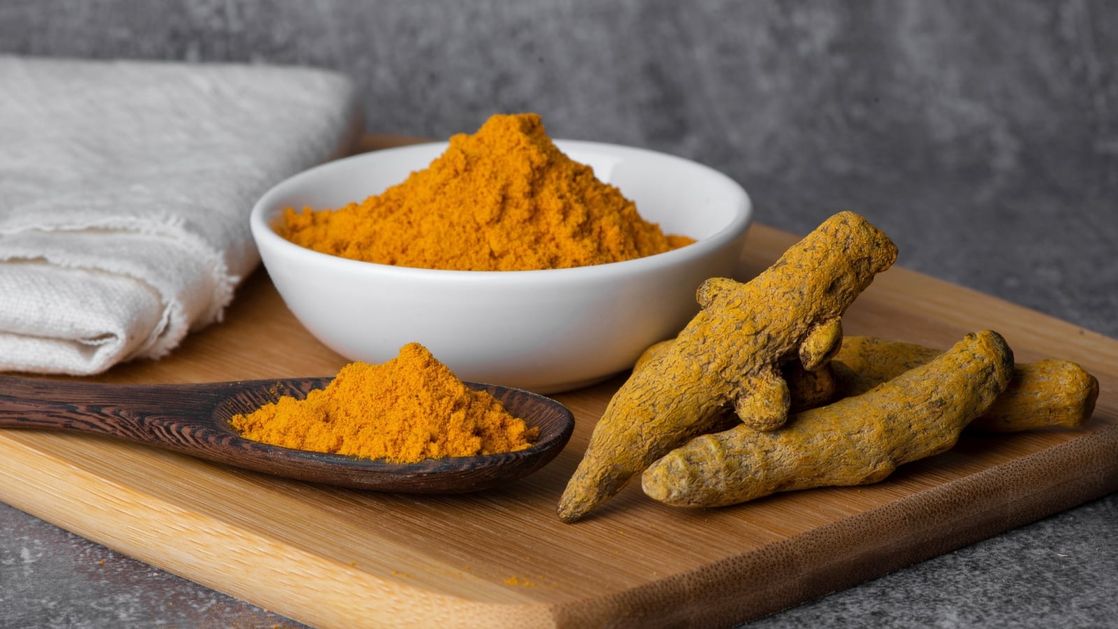 Bright yellow turmeric powder rests on a white plate, with fresh turmeric roots placed on a cutting board beside white fabric.