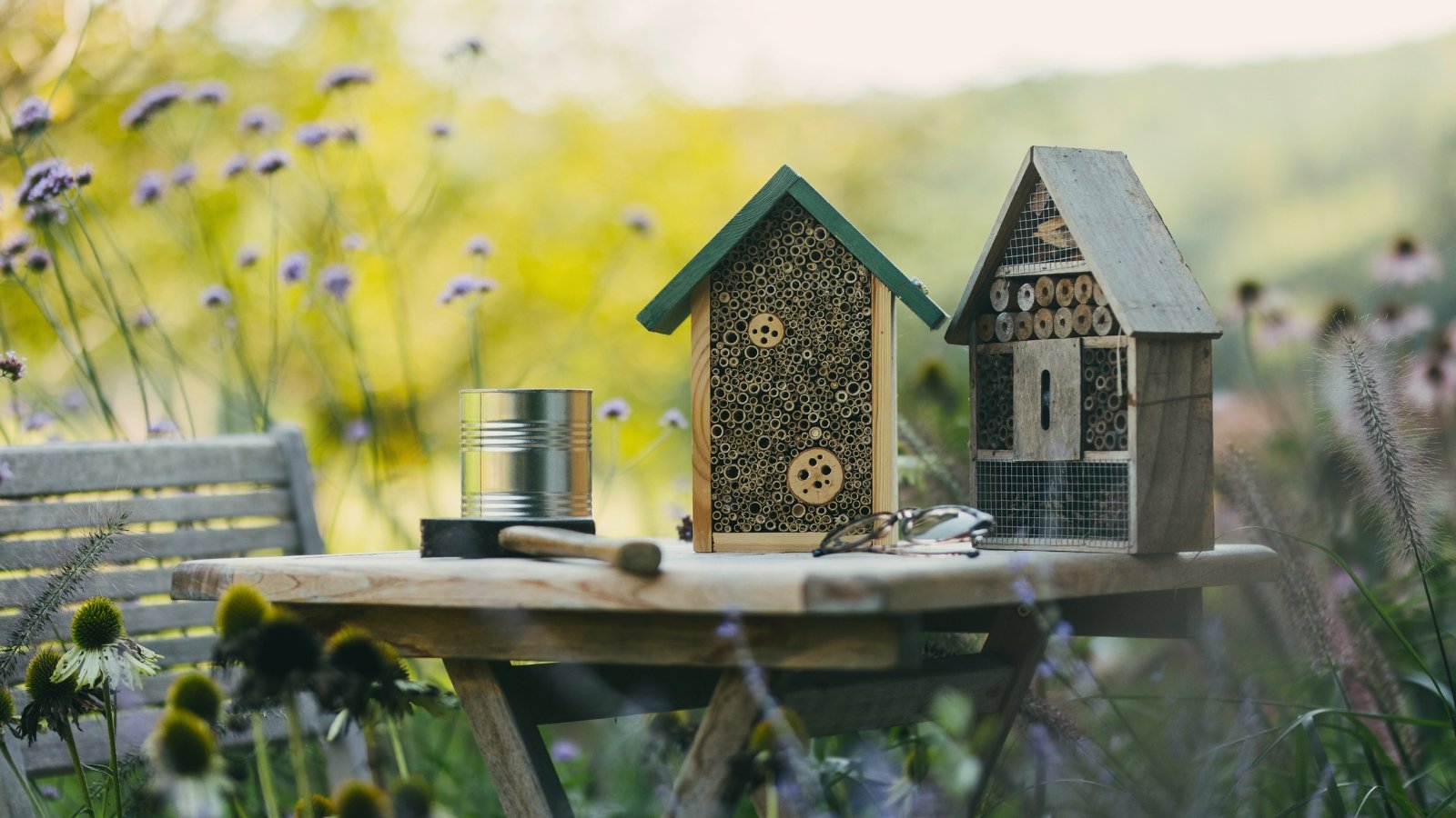 Two wooden insect houses on a table in a blooming garden, surrounded by a hammer, glasses, and a small metal barrel.