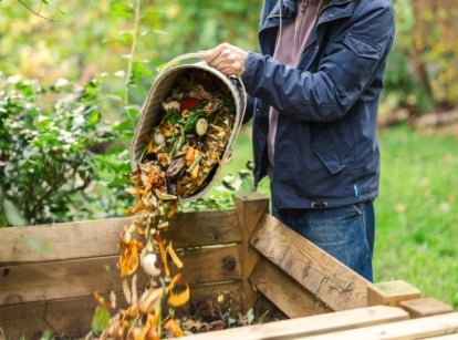 A gardener tosses fresh vegetable scraps into a compost container in the garden, illustrating practical fall compost tips for enriching soil.
