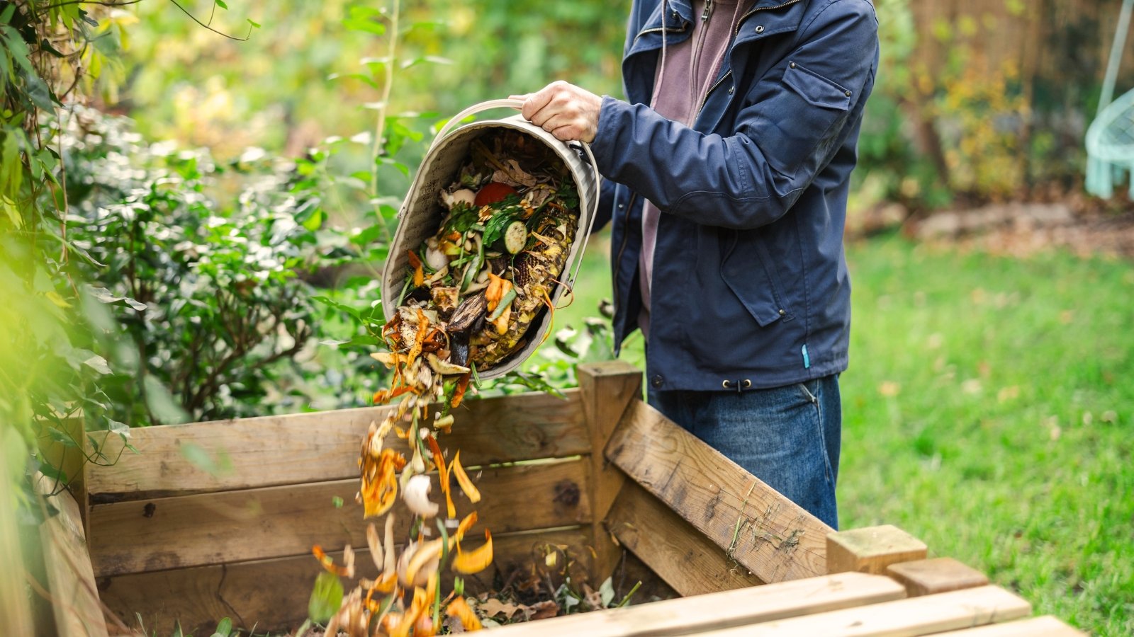 A gardener tosses fresh vegetable scraps into a material container in the garden, illustrating practical fall compost tips for enriching soil.