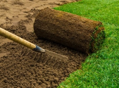 A gardener rakes the loose brown soil mix in a garden bed, preparing the surface before rolling out a layer of new green lawn.