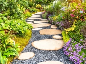 A neat rock path free of weeds winds through a garden, bordered on one side by lush woodland and tropical plants and on the other by blooming perennials, native greenery, and low-growing groundcovers.
