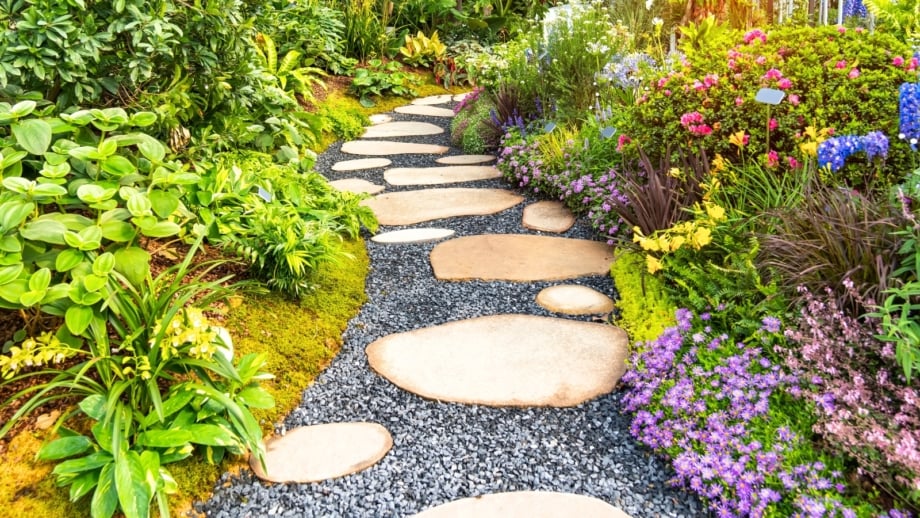 A neat rock path free of weeds winds through a garden, bordered on one side by lush woodland and tropical plants and on the other by blooming perennials, native greenery, and low-growing groundcovers.