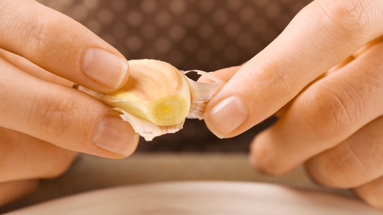 A close-up shot of a person's hand in the process of peeling a small clove of an allium bulb, all situated in a well lit area