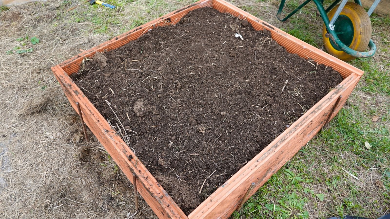 An overhead and close-up shot of a small raised bed filled with rich brown soil, with a wheelbarrow right beside it