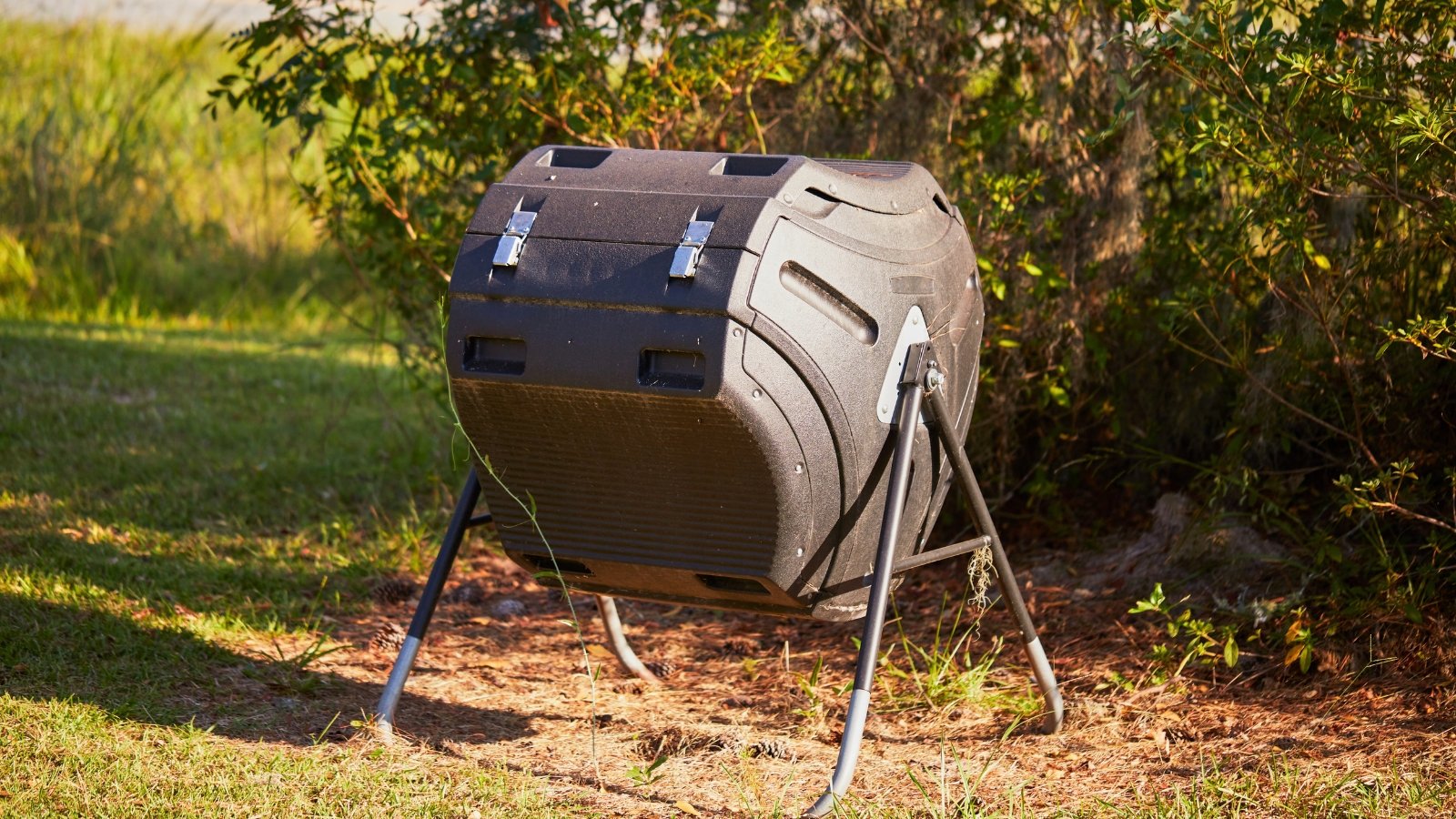 A black compost tumbler stands in the garden, its cylindrical body elevated on a frame for easy rotation and aeration of the organic material inside.
