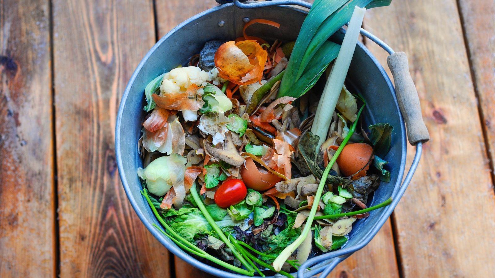 Top view of a bucket filled with organic waste, such as fruit peels and vegetable scraps, placed on a wooden floor.
