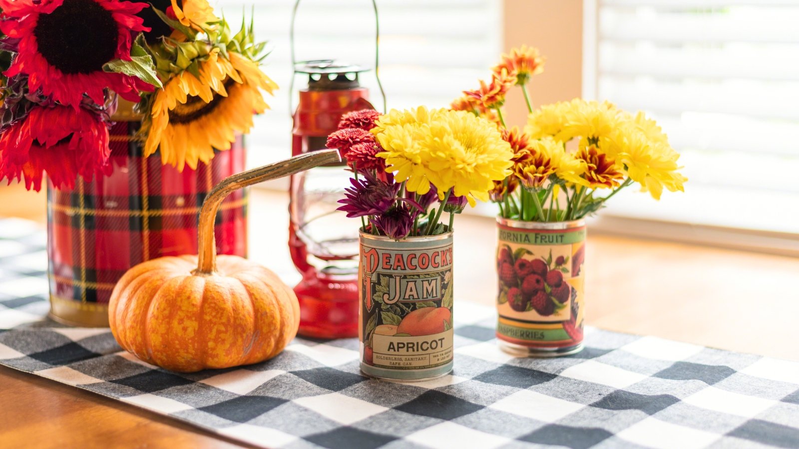 A plaid-covered container and vintage tin cans are used as vases for sunflowers, chrysanthemums, and red celosia, placed next to a small gourd.