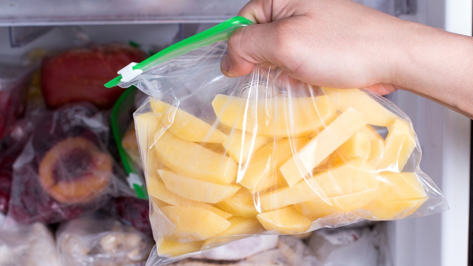 A hand holds a zip-lock bag filled with sliced frozen potatoes against a freezer stocked with various frozen fruits.