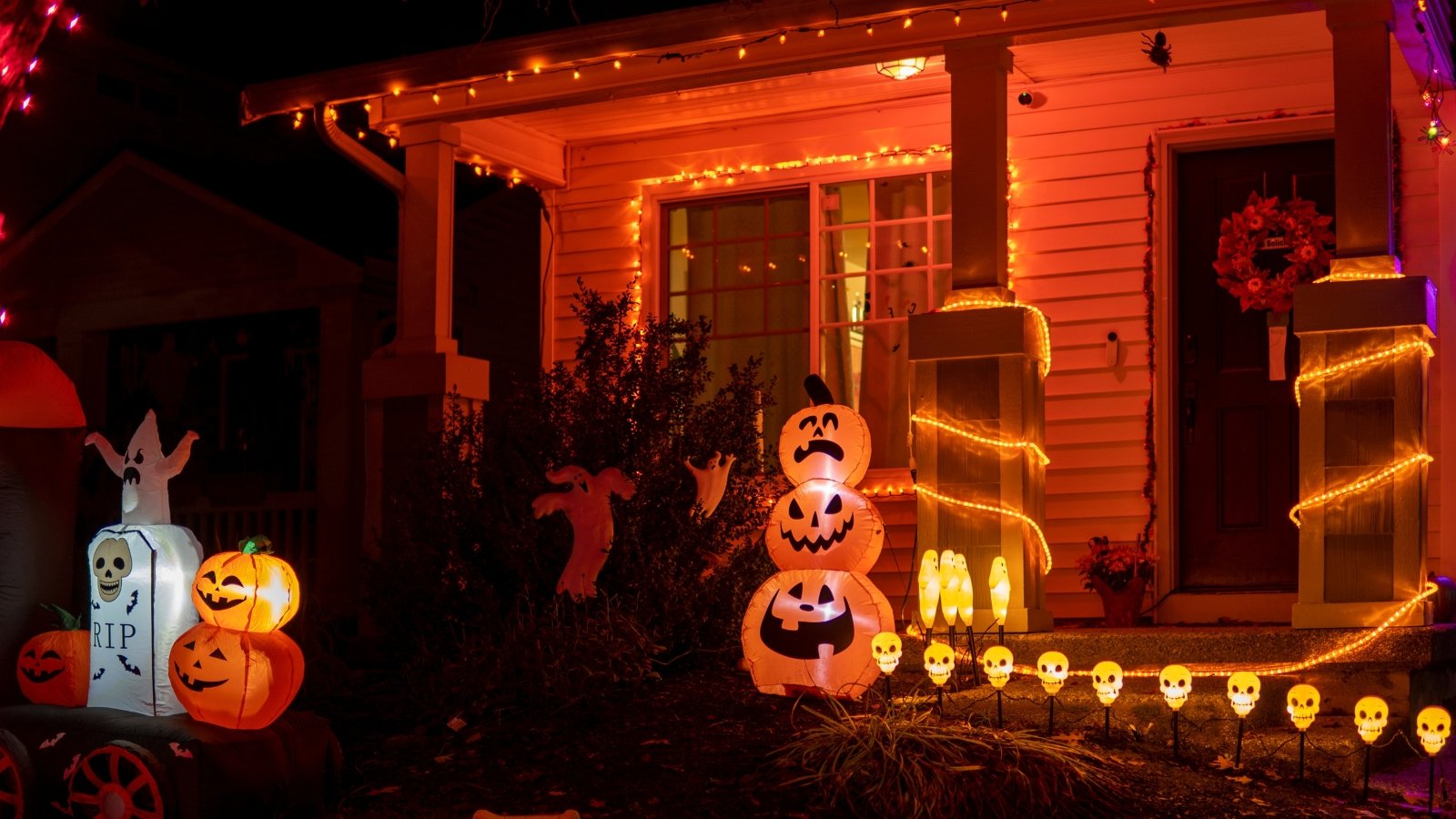 Glowing Halloween outdoor decorations light up the house porch with carved pumpkins, ghost figures, and orange garlands creating a festive, spooky atmosphere.