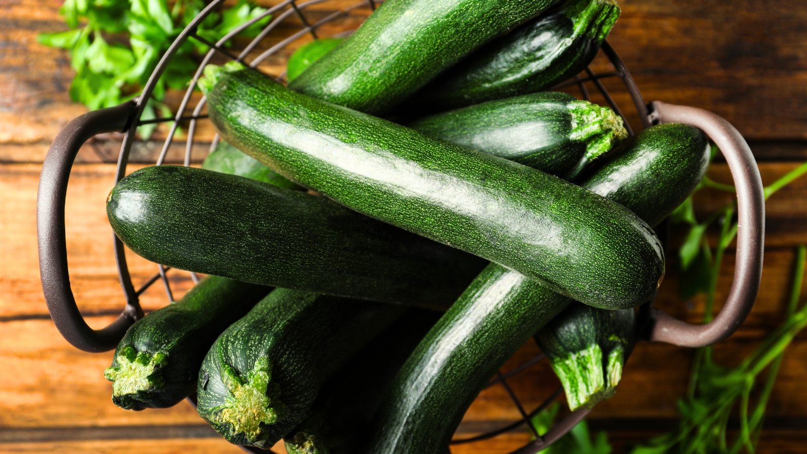 Several dark, glossy squash fruits fill a basket resting on a wooden table, their smooth surfaces reflecting light.