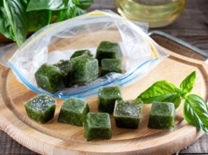 Ice cubes with frozen basil leaves glisten on a wooden table beside fresh sprigs, showing an easy way to freeze garden herbs for later use.