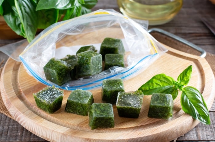 Ice cubes with frozen basil leaves glisten on a wooden table beside fresh sprigs, showing an easy way to freeze garden herbs for later use.