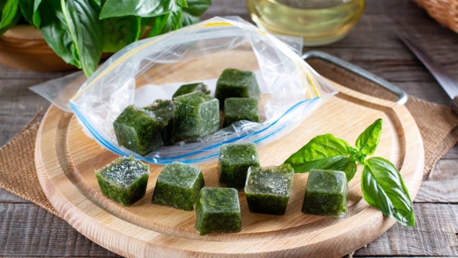 Ice cubes with frozen basil leaves glisten on a wooden table beside fresh sprigs, showing an easy way to freeze garden herbs for later use.