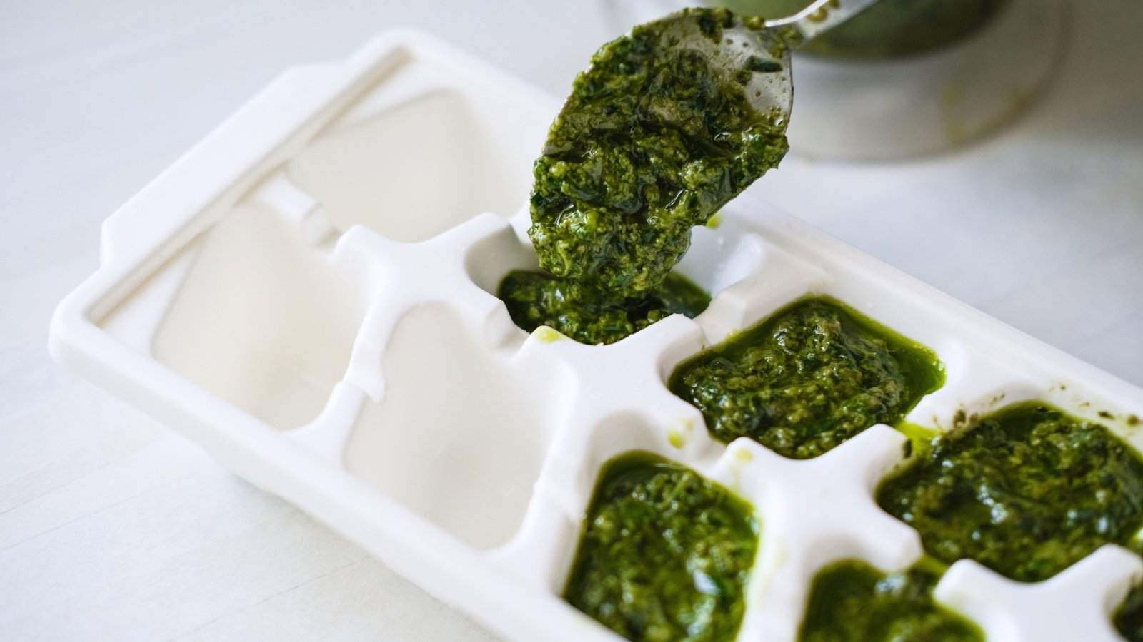 A white ice cube tray is being filled with vibrant green pesto using a spoon, ready for freezing.
