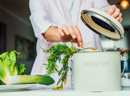 Indoor composting scene with a woman adding vegetable peelings to a small countertop compost bin, where carrot leaf scraps hang over the edge.