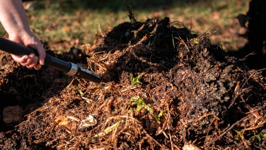A gardener uses a garden pitchfork to turn a steaming heap of organic matter in an autumn setting, illustrating october composting in progress.