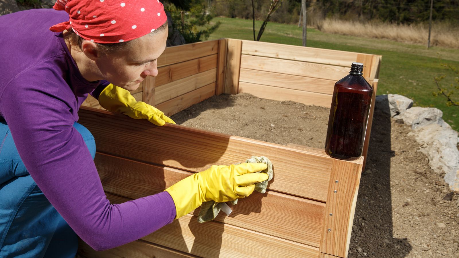 A close-up shot of a person in the process of applying oil onto a large wooden planter, all situated in a large garden area