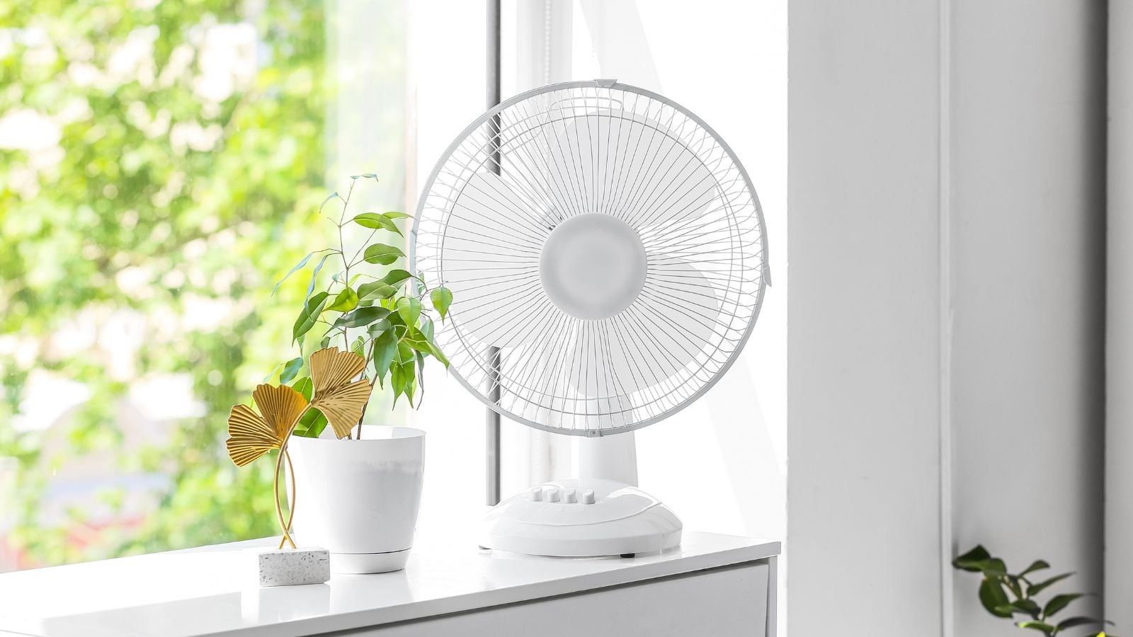 A close-up shot of a small white colored ventilator, placed beside a potted houseplant on a windowsill indoors