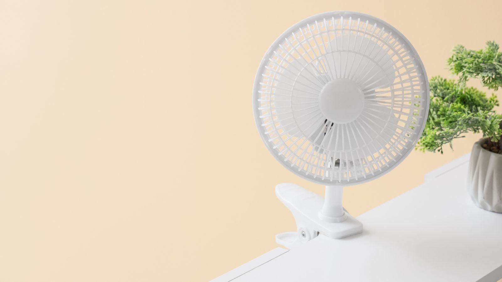 An overhead and close-up shot of a small clip-on desk ventilator, placed beside a small potted houseplant, in a well lit area