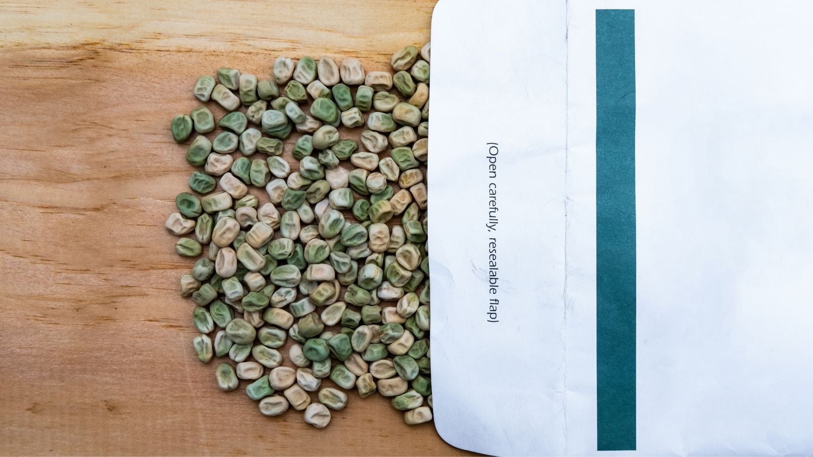 A close-up and overhead shot of a pile of dried seeds of a crop, pouring out from a paper packet, placed on a wooden surface