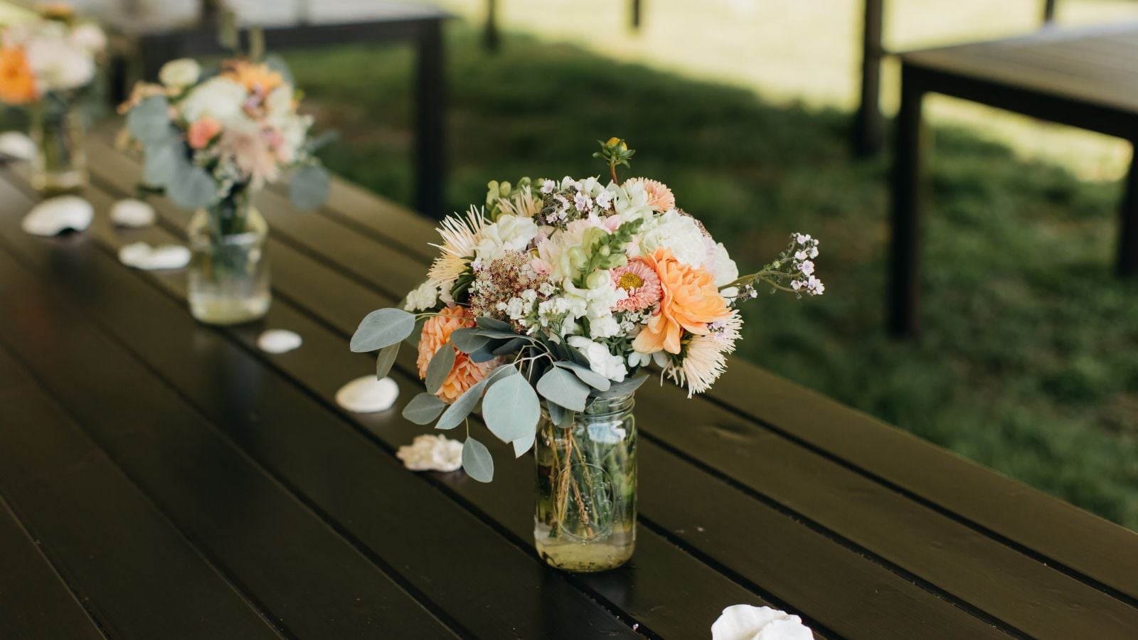 A close-up shot of a simple bloom arrangement in a jar, all arranged in rows, on top of a wooden table in a well lit area outdoors