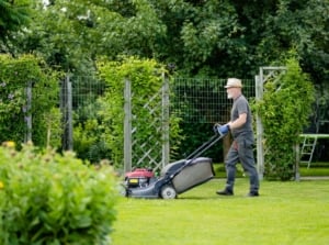 A man working to avoid spring lawn mistakes, using a lawn mower to trim the are surrounded by other bushes and trees