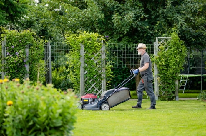 A man working to avoid spring lawn mistakes, using a lawn mower to trim the are surrounded by other bushes and trees