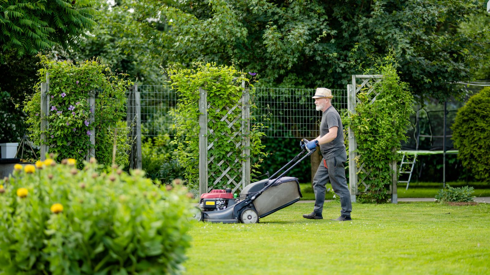 A man working to avoid spring lawn mistakes, using a lawn mower to trim the are surrounded by other bushes and trees