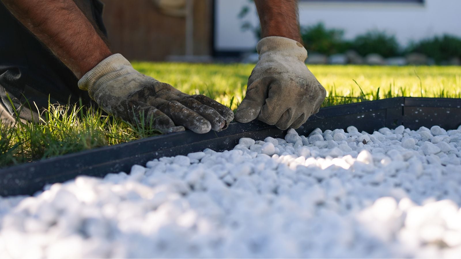 A close-up shot of a person's hands, wearing dirty gloves, in the process of installing a metal border that separates the lawn and a pebbled surface