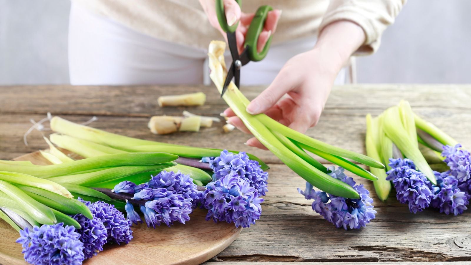 A woman using scissors to cut stems of a blooming plant, laying the pieces on a worn and wooden surface