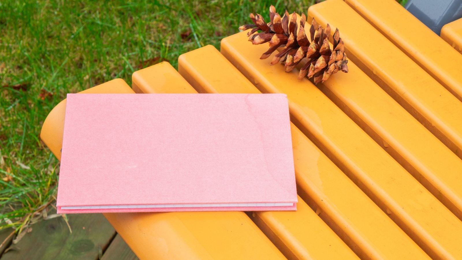 A close-up and overhead shot of a pink, unlined notebook or Garden Journal, placed on a yellow colored metal bench alongside an acorn outdoors