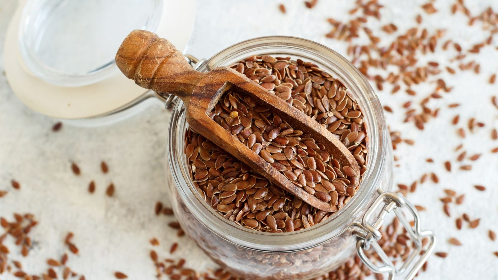 A close-up and overhead shot of a glass mason jar filled with small brown ovules and a wooden scooper, all palced on a white surface indoors